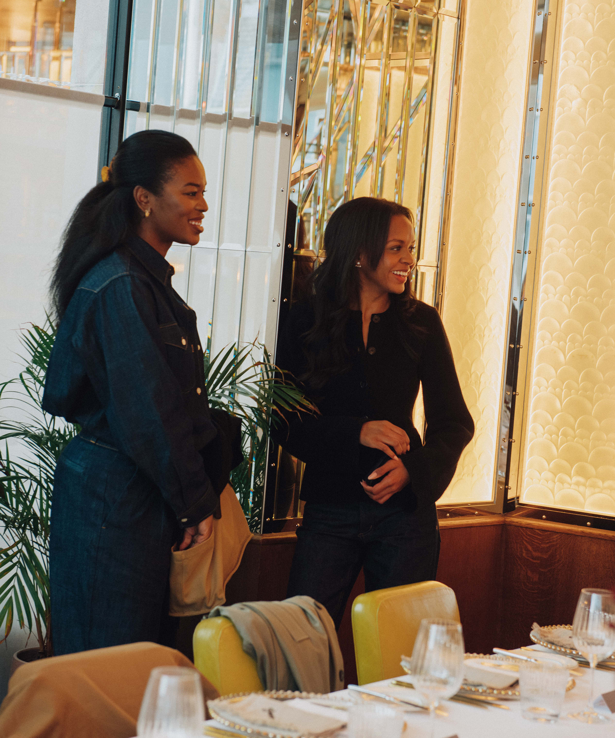 Two women stand and smile beside a table set for a meal in Selfridges’ stylish, warmly lit restaurant, featuring yellow chairs, mirrored walls, and decorative plates and glasses on the table.