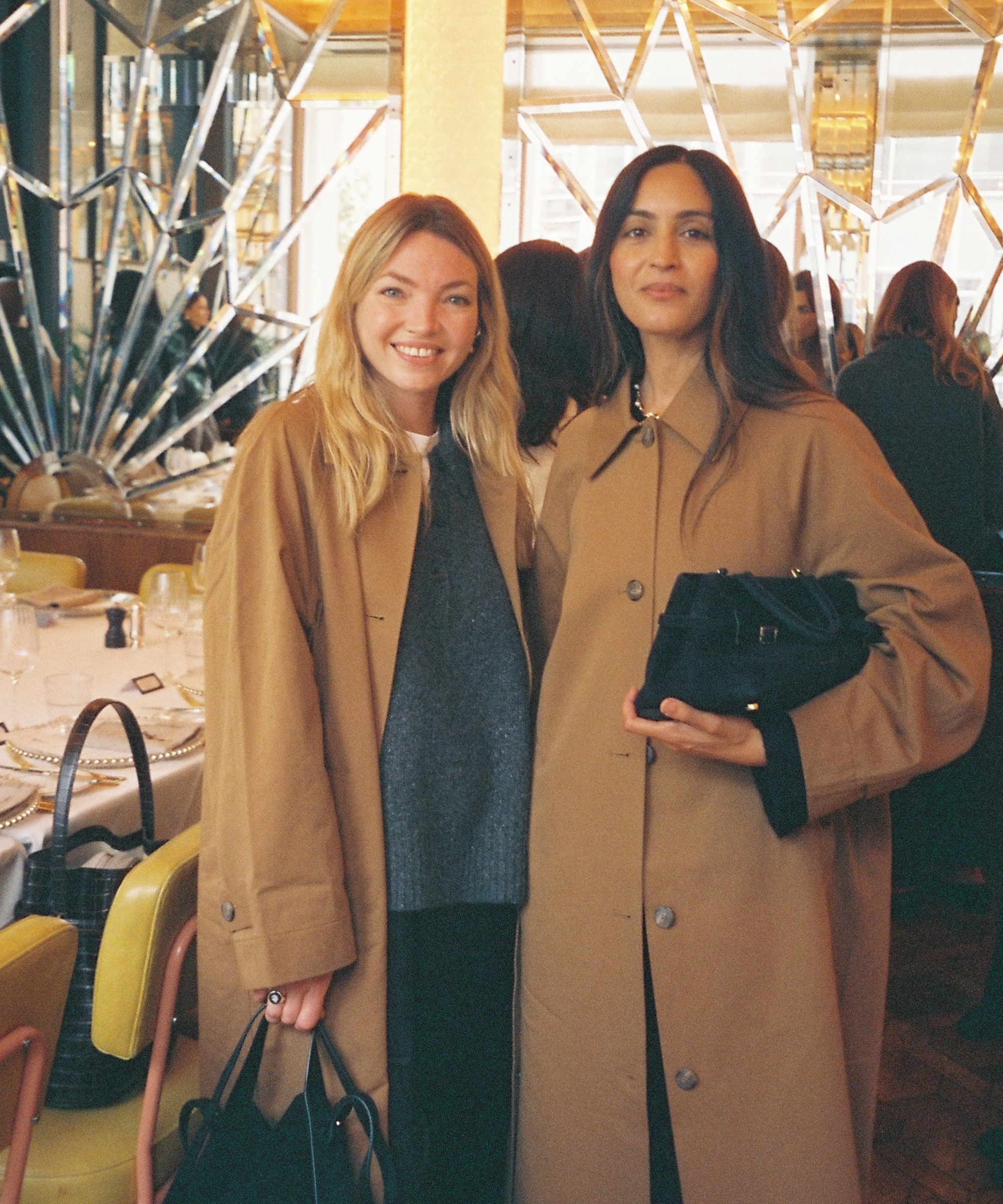 Two women stand side by side indoors at Selfridges, both wearing tan coats. One has blonde hair and smiles at the camera, while the other has long dark hair and holds a black handbag. Dining tables and people are visible in the background.