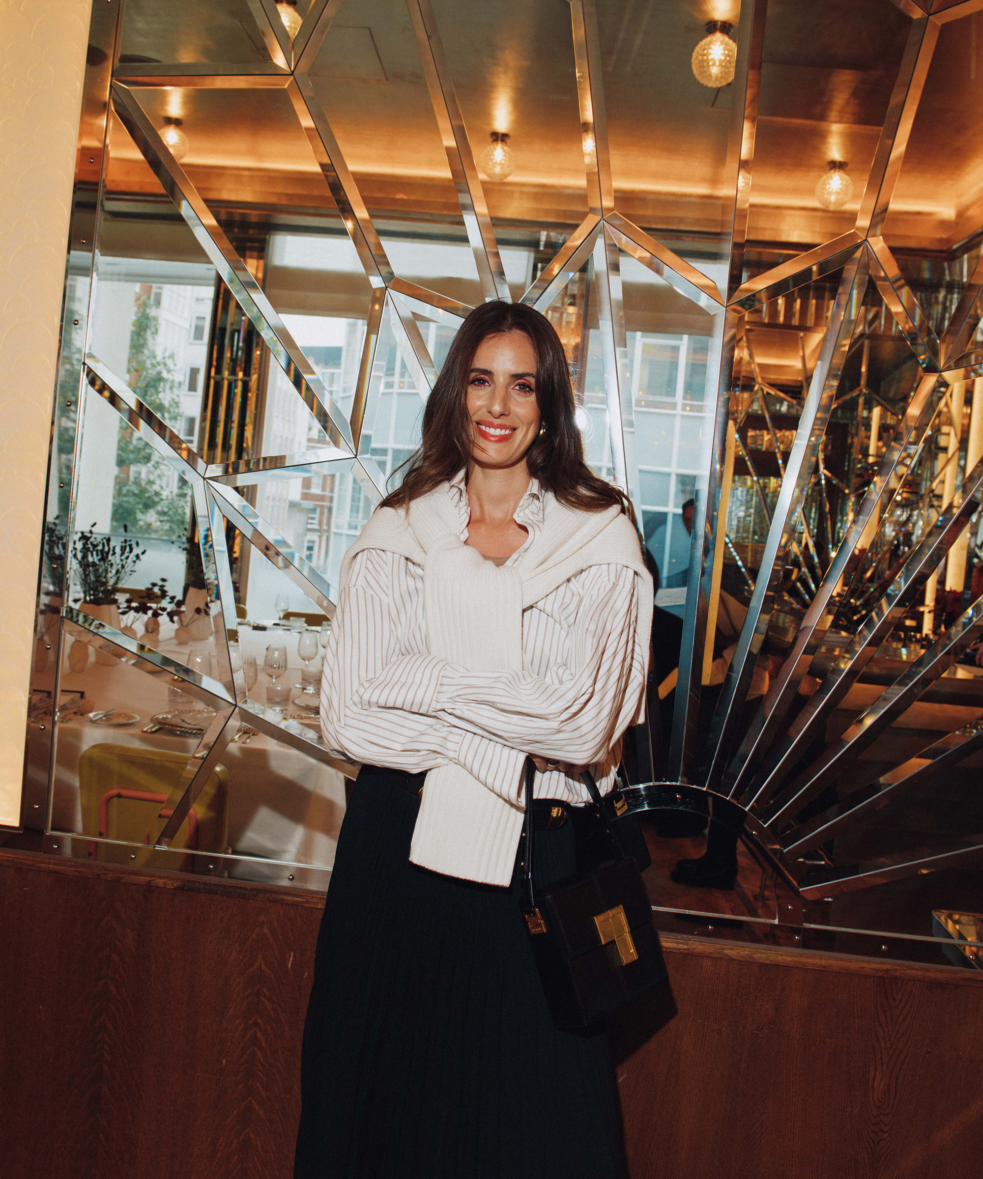 A woman with long brown hair, wearing a white sweater over a striped shirt and a black skirt, stands smiling in front of a decorative mirrored wall in a stylish Selfridges restaurant.
