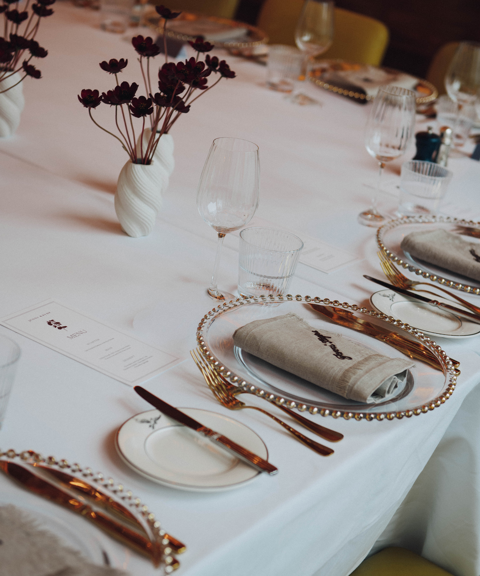 A formal dining table set with elegant plates, gold cutlery, glasses, light gray napkins, and decorative pearl-rimmed chargers from Selfridges. Minimalist vases with dark flowers are arranged on a crisp white tablecloth.