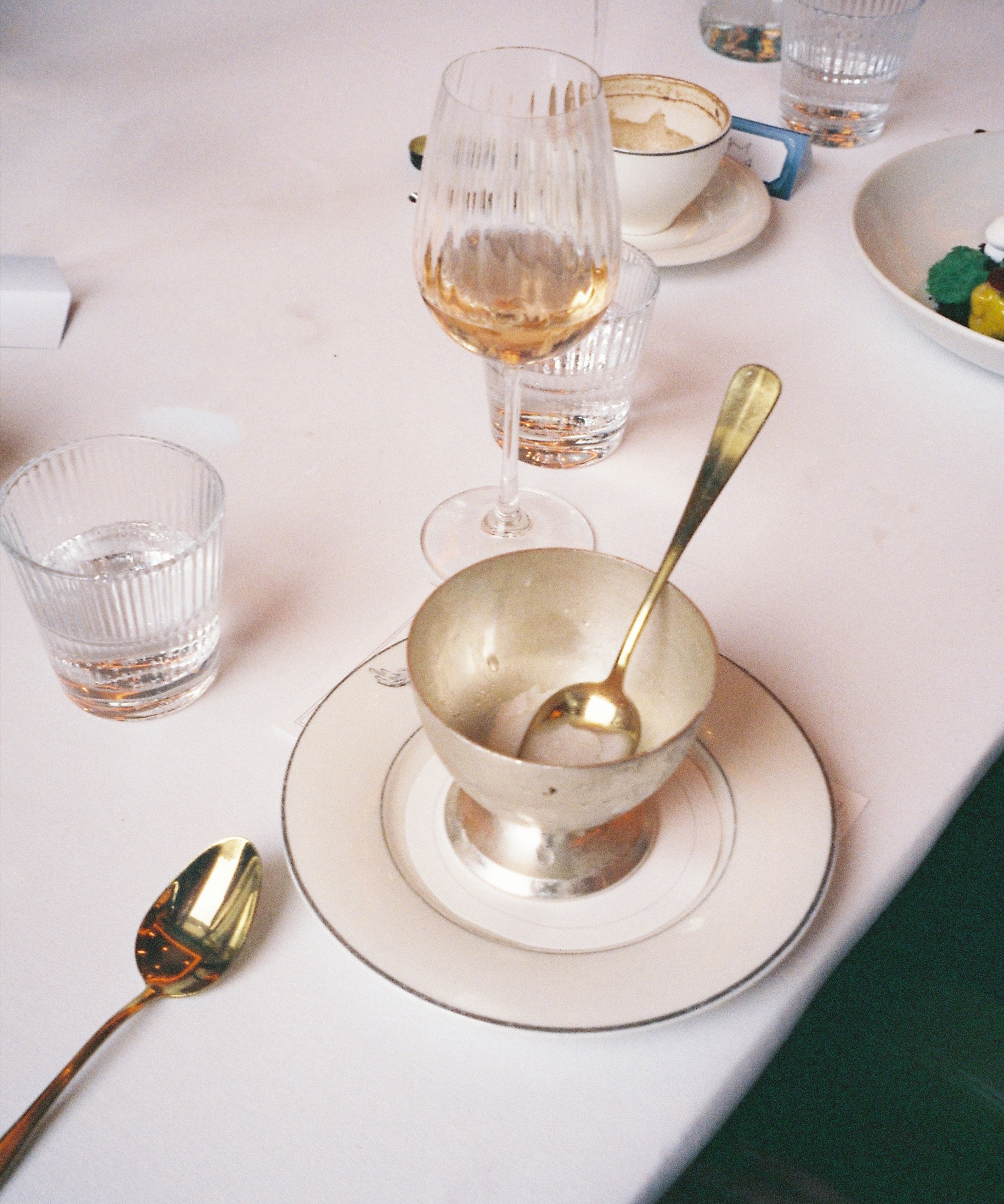 A white tablecloth holds a glass of white wine, a clear water glass, an empty silver dessert cup with a spoon on a plate, another spoon beside it, and a white cup and saucer in the background at Selfridges.