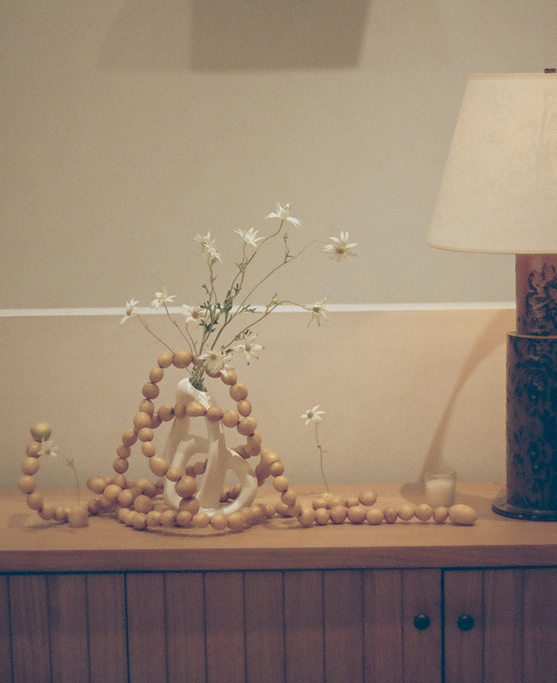 A wooden sideboard with a white sculptural vase holding delicate white flowers, draped with a long garland of small tan beads—perfect Friendsgiving decor—next to a table lamp and a small candle.