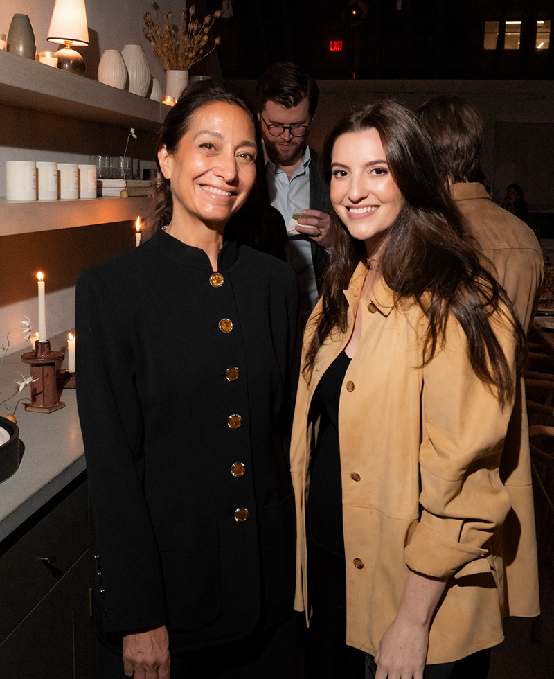 Two women stand smiling in a warmly lit room at a friendsgiving, wearing dark and tan jackets. The background shows candles, shelves with decorative items, and people engaged in conversation.