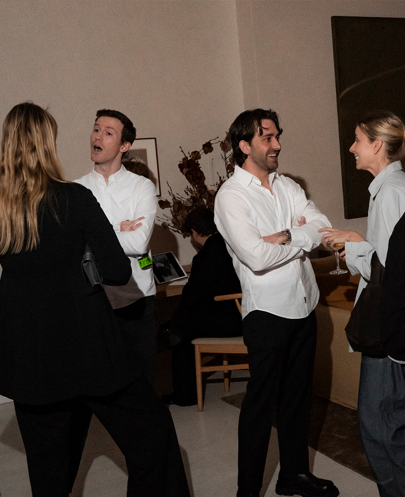 Four people socializing indoors at a friendsgiving event; two men in white shirts chat with two women, one in black and one in gray, while a person sits at a table in the background.