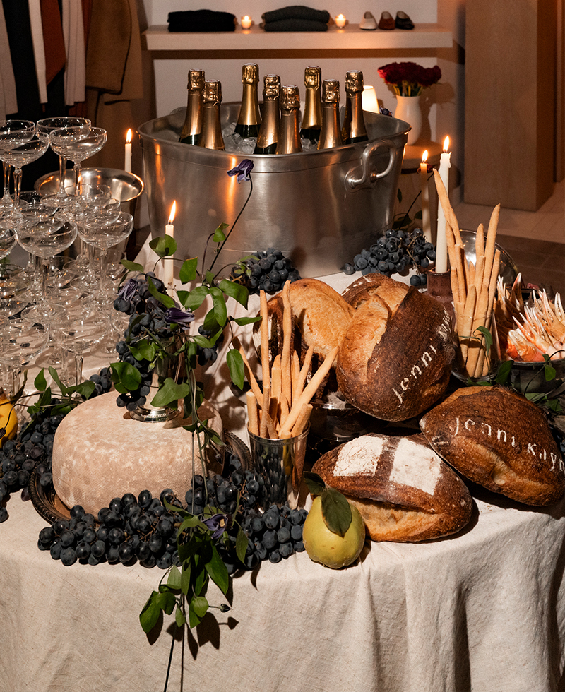 A table covered with a white cloth is set for friendsgiving, featuring loaves of bread, cheese, grapes, and foliage. Champagne bottles chill nearby with stacked glasses, while lit candles create a warm ambiance.