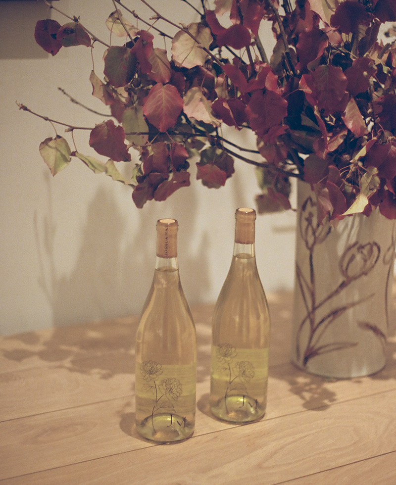 Two bottles of white wine with floral labels sit on a wooden table next to a large vase filled with reddish-brown and green leaves, creating an inviting scene perfect for a cozy Friendsgiving gathering.