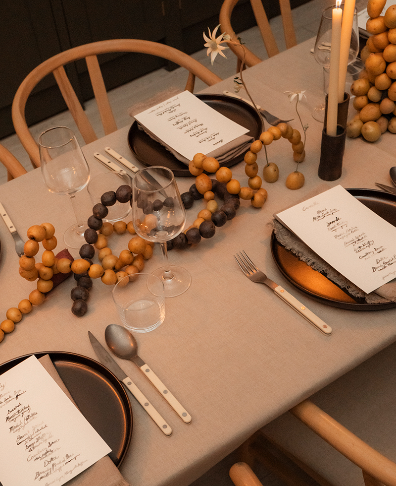 A neatly set dining table with beige linens, black plates, white-handled cutlery, wine glasses, menus, candles, and decorative wooden bead garlands—perfect for a warm Friendsgiving gathering. Light wood chairs surround the table.