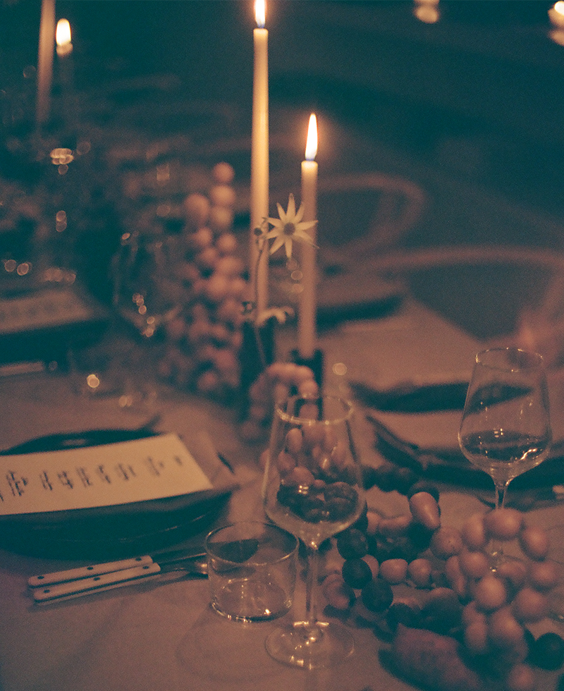 A dimly lit dinner table set with wine glasses, plates, cutlery, candles, and a decorative garland creates a warm and elegant Friendsgiving atmosphere.
