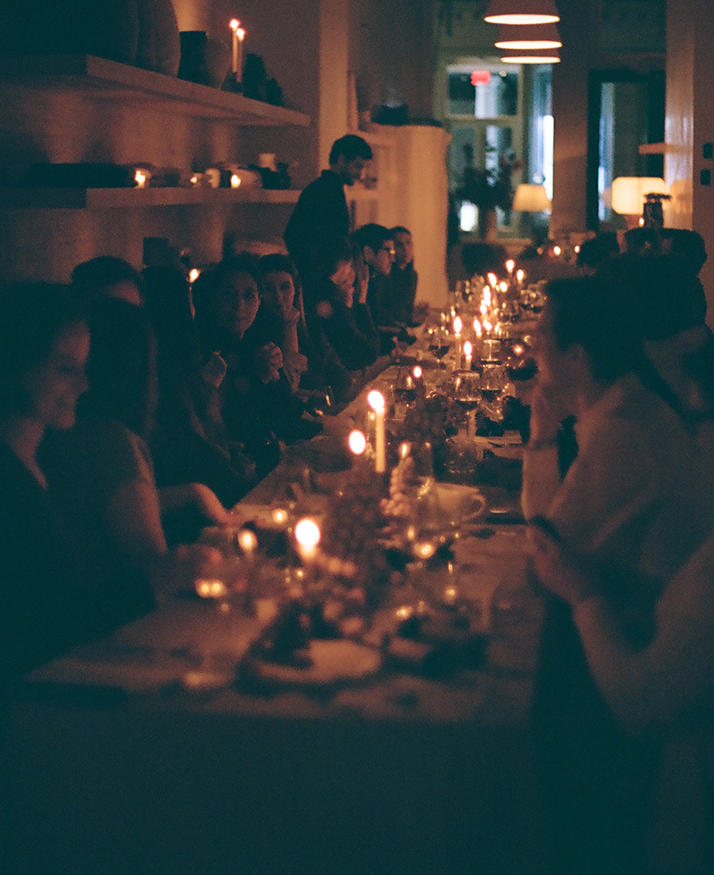A dimly lit, elegant Friendsgiving dinner party with many people seated at a long table, illuminated by candlelight. The atmosphere is warm and intimate, with soft shadows and cozy interior decor visible in the background.