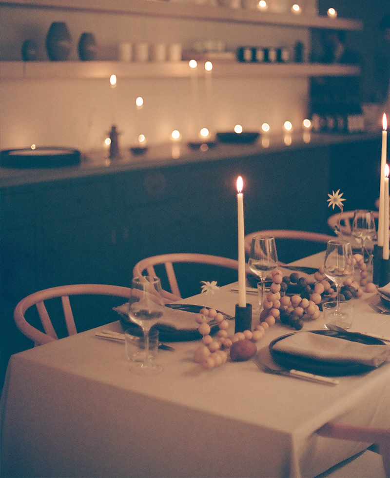 A softly lit dining table with a white tablecloth is set for Friendsgiving, featuring plates, glasses, and cutlery. Tall candles and a garland of wooden beads decorate the table; shelves with more candles glow in the background.