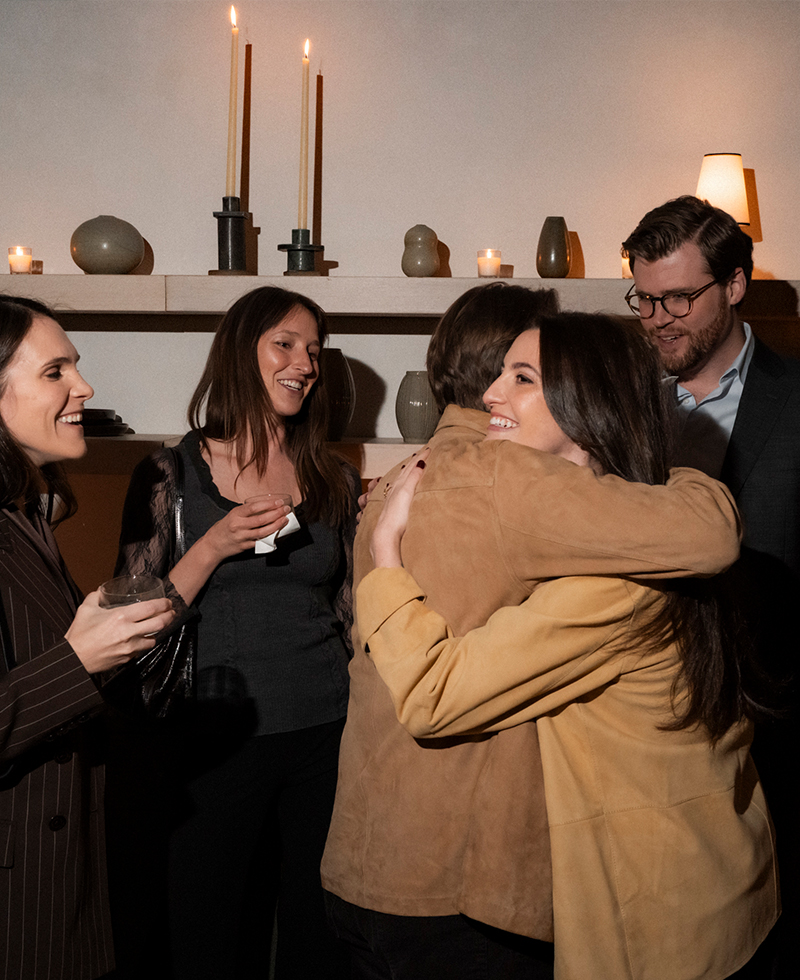 A group of five people at a warmly lit friendsgiving gathering, smiling and talking. Two people in the center embrace in a hug while others stand nearby holding drinks. Candles and ceramics decorate the shelves behind them.