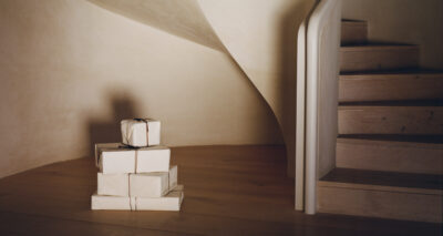 A stack of six neatly wrapped gift boxes tied with string sits on a wooden floor next to a curved staircase with light-colored wooden steps and a smooth, cream-colored wall.