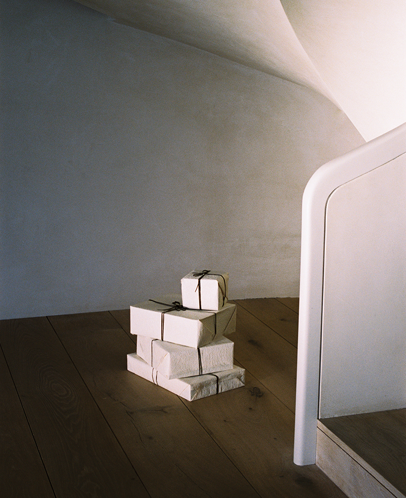 A stack of small, neatly wrapped parcels tied with string sits on a wooden floor beside a pale, curved staircase against a minimal, light-colored wall.