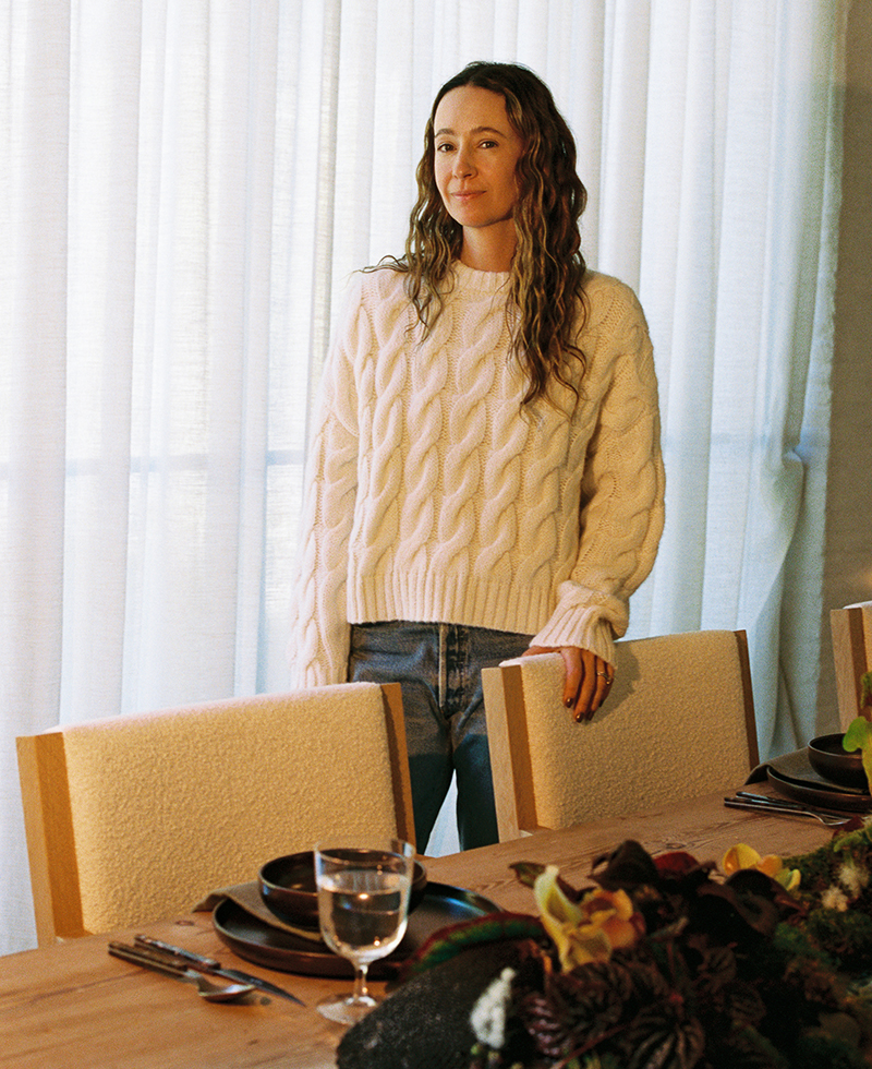 A woman with long wavy hair, wearing a white cable-knit sweater and jeans, stands beside a set dining table with plates, glasses, and a floral centerpiece, in front of sheer white curtains.