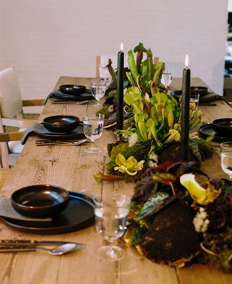 A wooden dining table set with black dishes, silver cutlery, clear glasses, and two lit black candles. A lush green and yellow floral centerpiece runs along the center of the table.