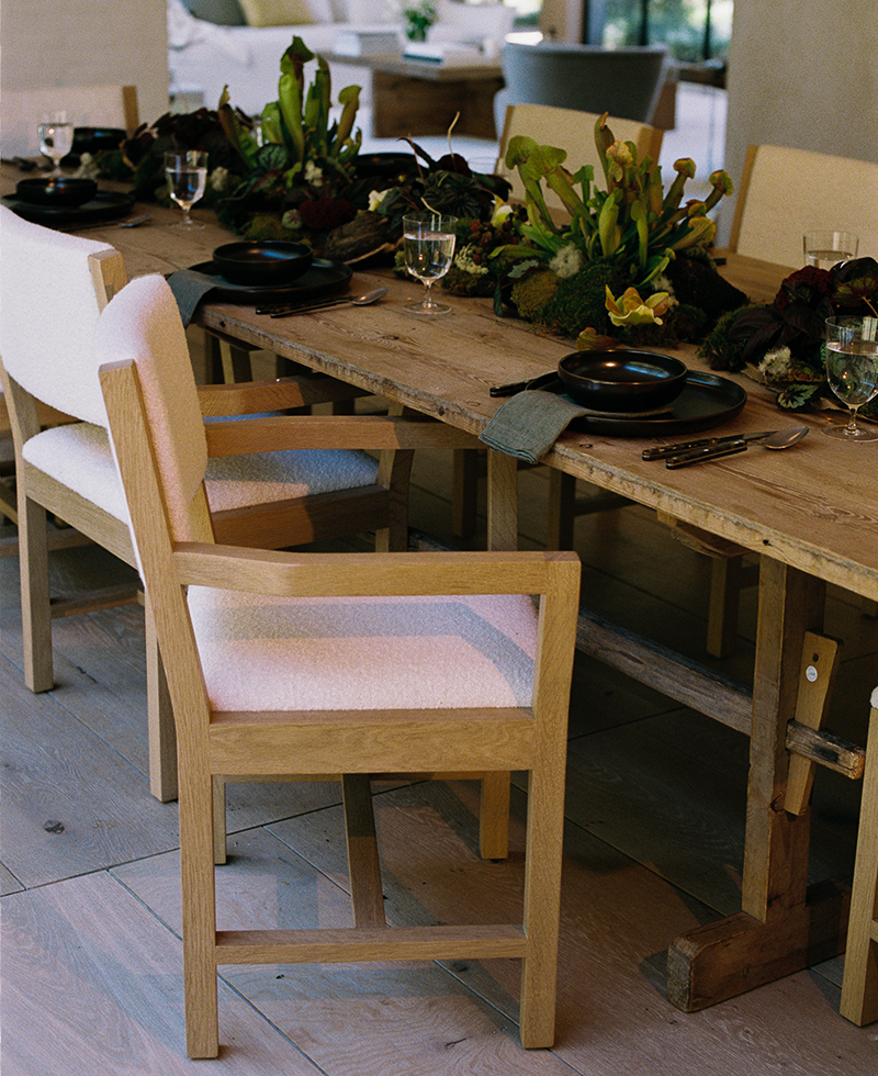 A rustic wooden dining table set with black plates, glassware, gray napkins, and a lush green centerpiece, surrounded by light wood chairs with white cushions. Sunlight filters into the cozy, inviting room.