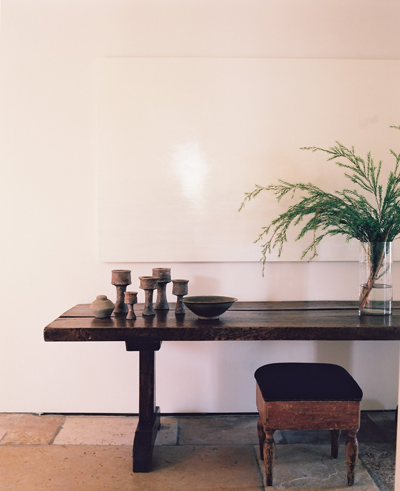 A rustic wooden table with candlesticks, a bowl, and a small vase sits against a white wall, evoking Tel Aviv's relaxed style. A glass vase with green foliage rests on the right side, while a simple stool is tucked underneath.