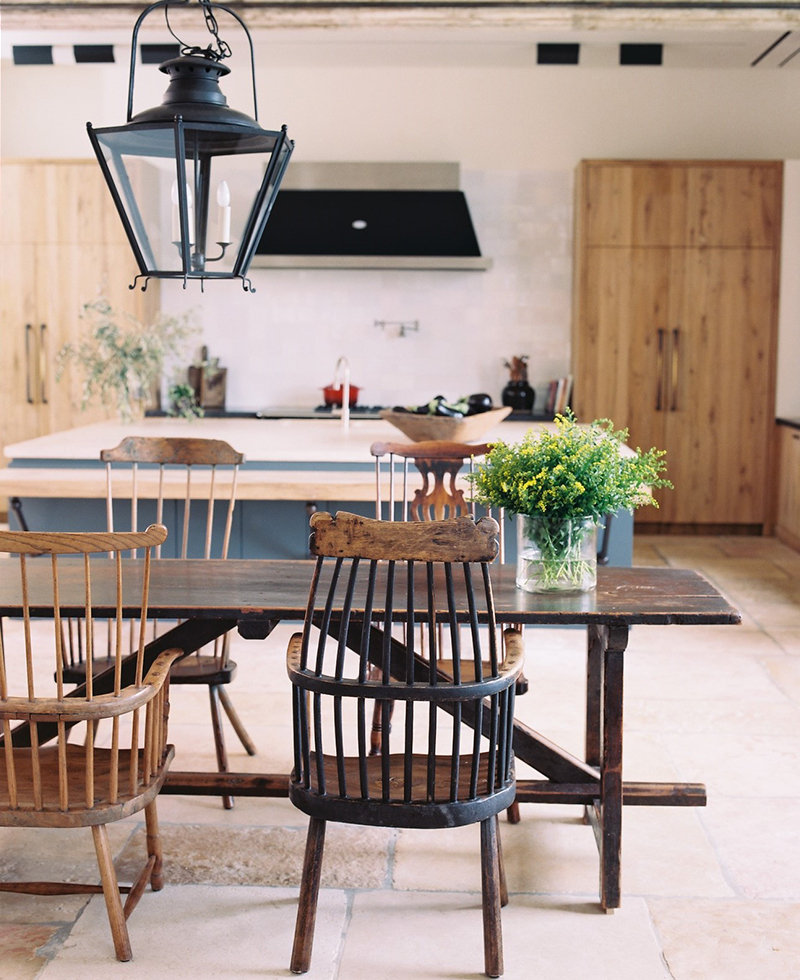 A rustic kitchen in Tel Aviv features a wooden dining table with mismatched chairs, a vase of yellow flowers, and a black lantern-style light fixture above. Light wood cabinets and a spacious island are visible in the background.