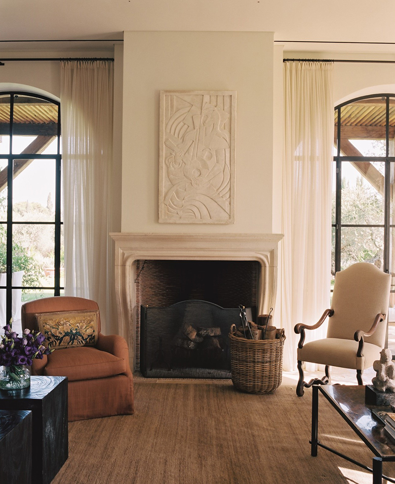 A cozy Tel Aviv living room with a fireplace and abstract art above it, featuring an armchair, a cushioned chair, sheer curtains, tall windows, a wicker basket, and a coffee table with flowers. Natural light fills the space.