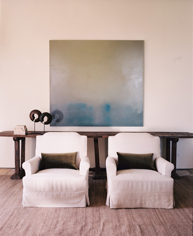 Two white armchairs with green accent pillows sit on a beige rug in front of a rustic wooden console table. Above, a large abstract painting in green and blue tones brings a touch of Tel Aviv style to the cream-colored wall.