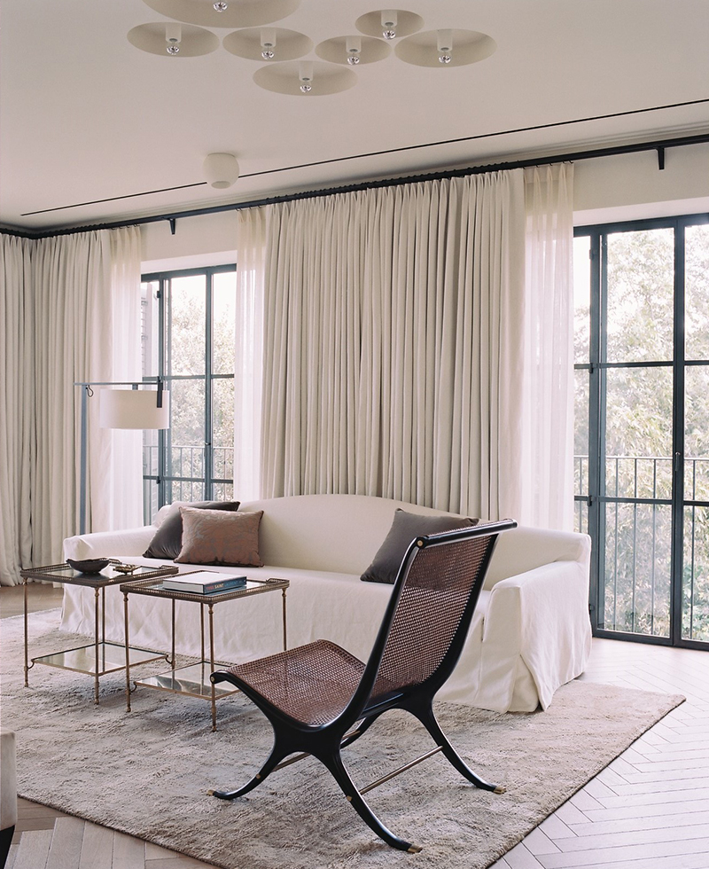 A modern Tel Aviv living room with large windows, sheer white curtains, a white sofa with cushions, a brown lounge chair, a glass coffee table, and a neutral rug on light wood floors.