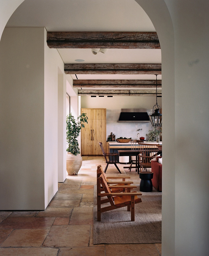 A cozy, rustic kitchen and dining area in Tel Aviv viewed through an archway, featuring wooden ceiling beams, natural stone floors, a wooden chair, a large potted plant, and a dining table with chairs.