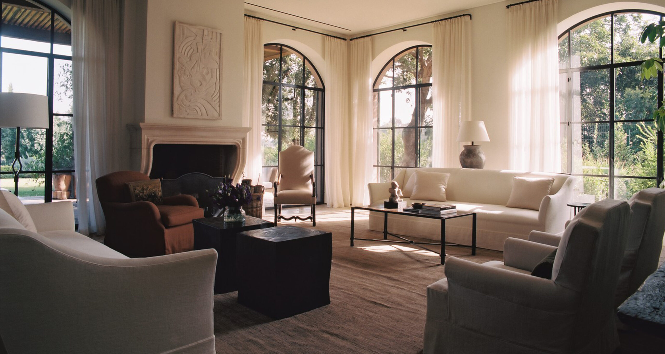 Elegant Tel Aviv living room with large arched windows, white sofas, armchairs, a fireplace, and natural light streaming in. Neutral tones and minimalist décor create a calm, inviting atmosphere.