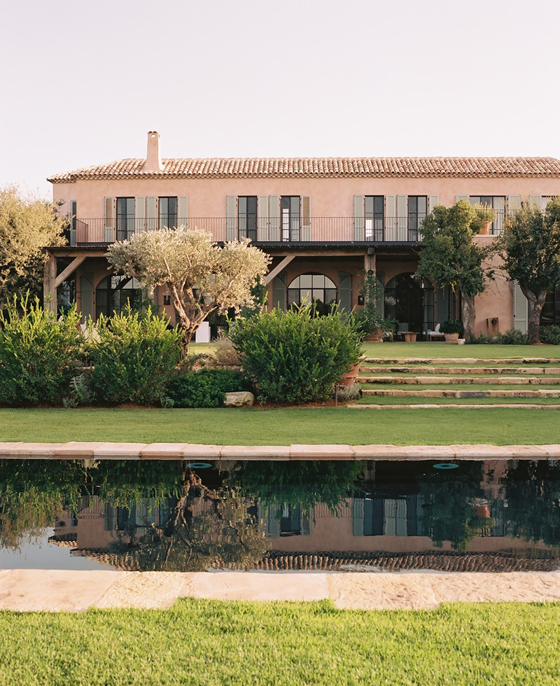 A large, two-story Mediterranean-style house in Tel Aviv with a tiled roof and arched windows, lush green lawns, steps, trimmed bushes, and a swimming pool in the foreground reflecting the home.