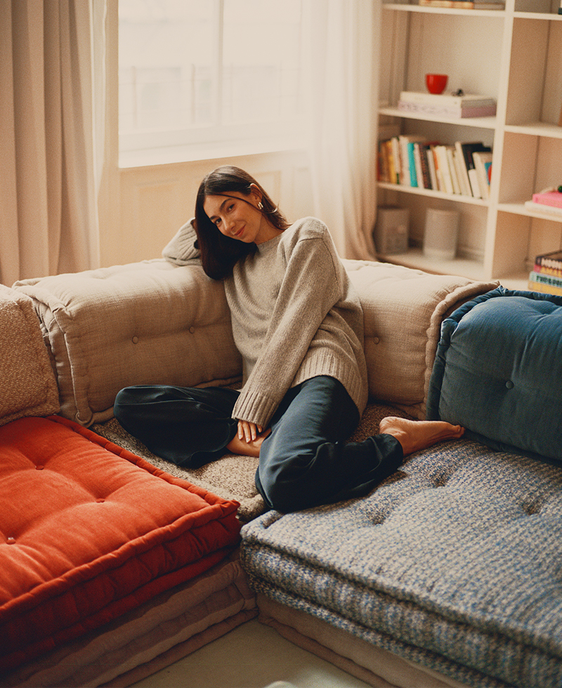 Mélanie Masarin, in a gray sweater and dark pants, sits barefoot and relaxed on a cozy sectional sofa with colorful cushions in a sunlit living room with bookshelves in the background.