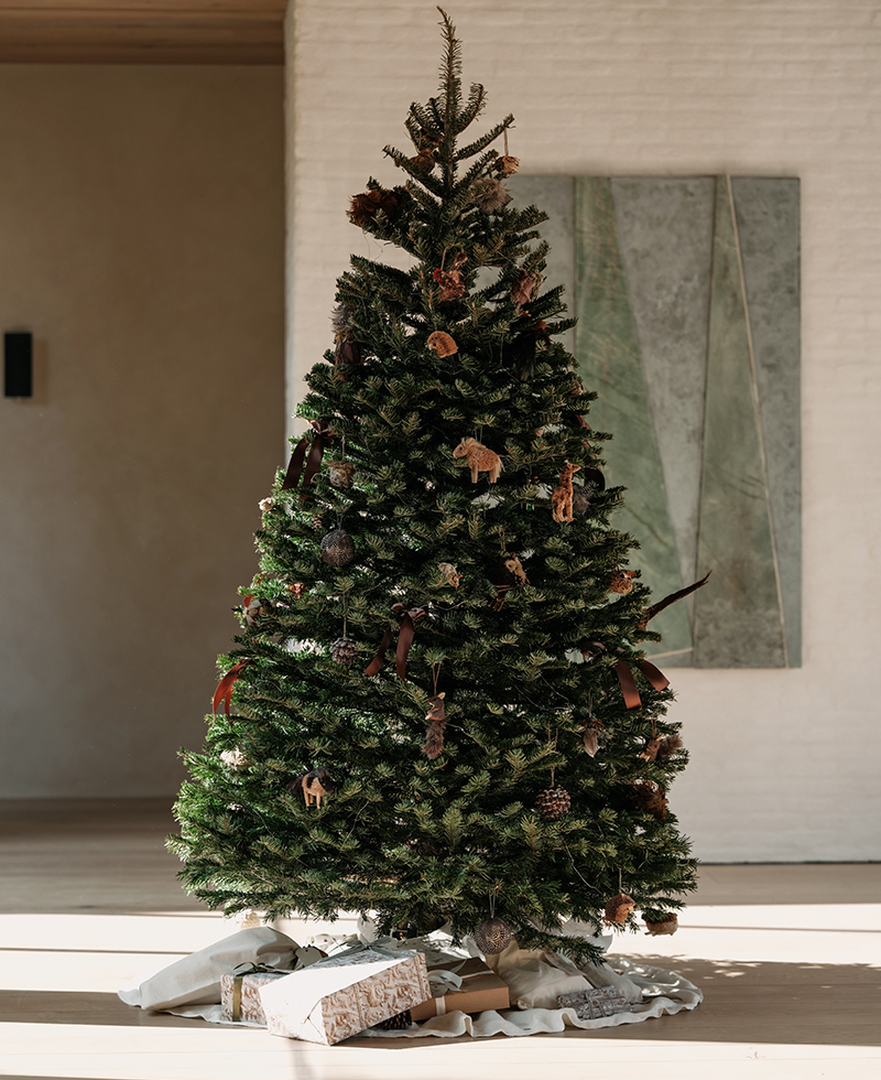A decorated Christmas tree stands indoors on a white tree skirt in this oak house, with a few wrapped presents underneath. The tree is adorned with rustic ornaments and ribbon, and a modern abstract painting hangs on the wall behind it.