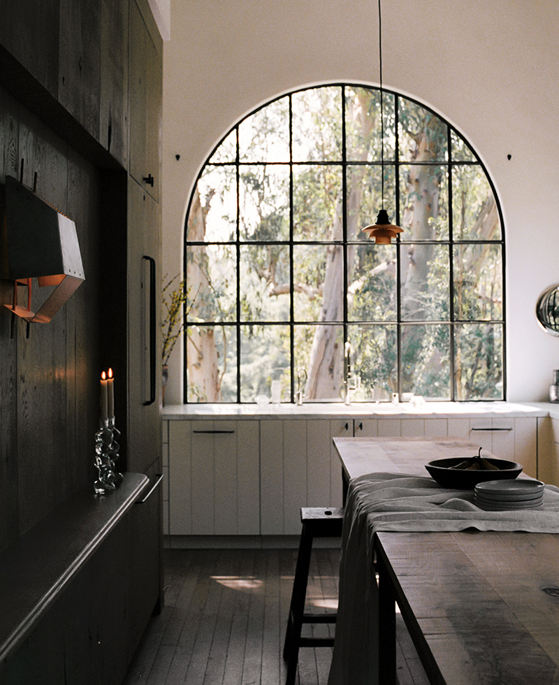 A modern kitchen with dark wood cabinets, a large arched window letting in natural light, white lower cabinets, a wooden island with a draped cloth, and two lit candles on a side shelf. Trees are visible outside the window.