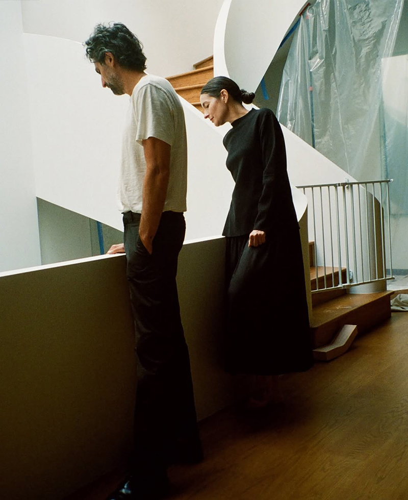 A husband in a white shirt and dark pants stands beside his wife in a long black dress by a white railing, both looking down thoughtfully in a modern, minimalist space filled with natural light.