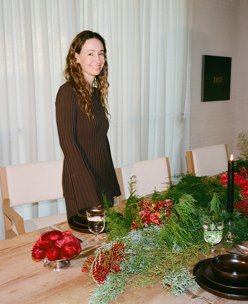 A woman with long, wavy hair in a dark dress stands smiling by a wooden table decorated with greenery, red flowers, black candles, and dinnerware in a softly lit room.