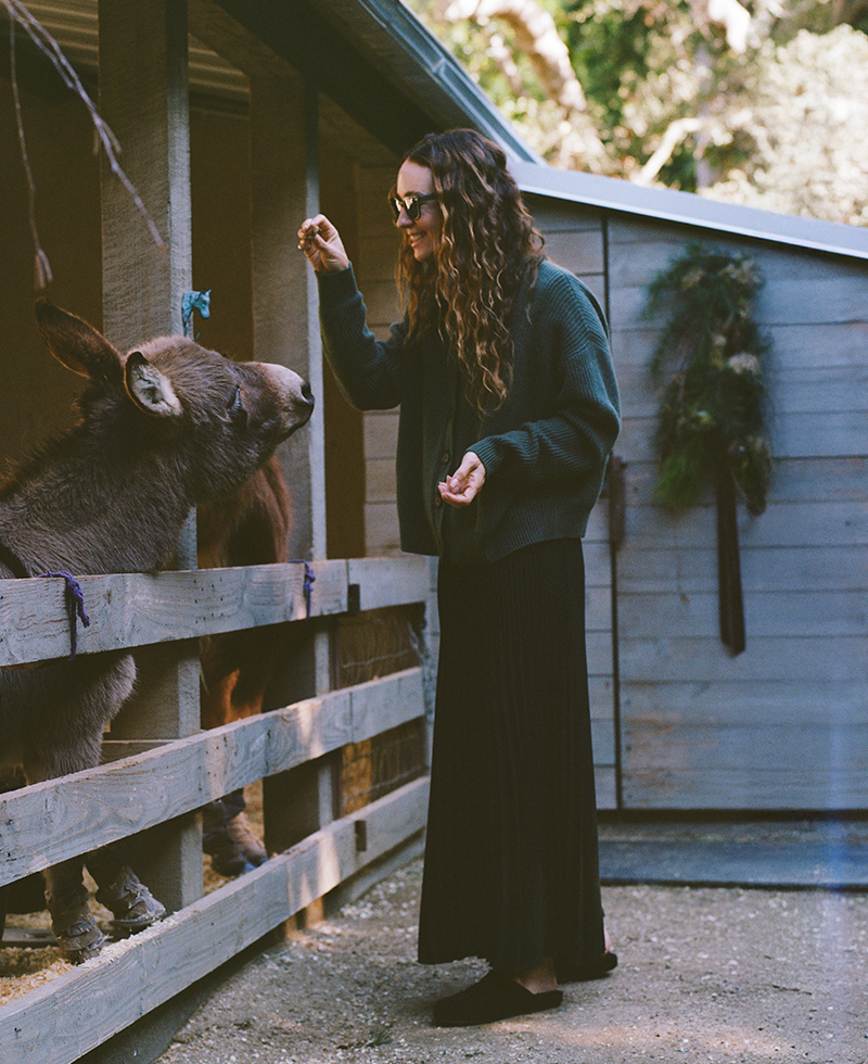 A woman with long curly hair, wearing sunglasses and a dark outfit, stands by a wooden fence, smiling and reaching out to feed a donkey outside a barn decorated with greenery.