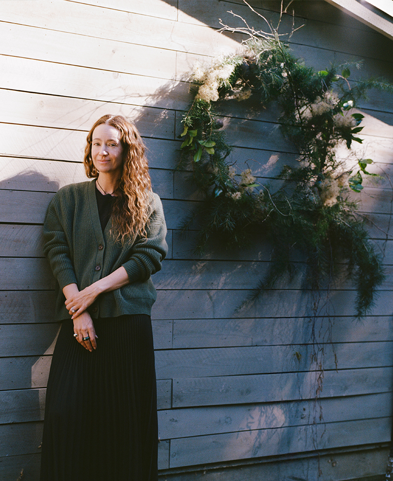 A woman with long curly hair, wearing a dark cardigan and skirt, stands in sunlight against a wooden wall decorated with a large wreath of greenery.