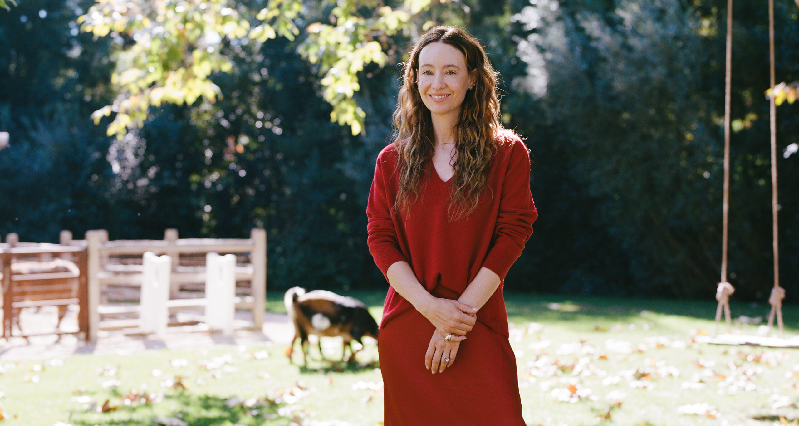 A woman in a long red dress stands smiling outdoors on a sunny day, with trees and a wooden fence in the background and a goat grazing on the grass nearby.