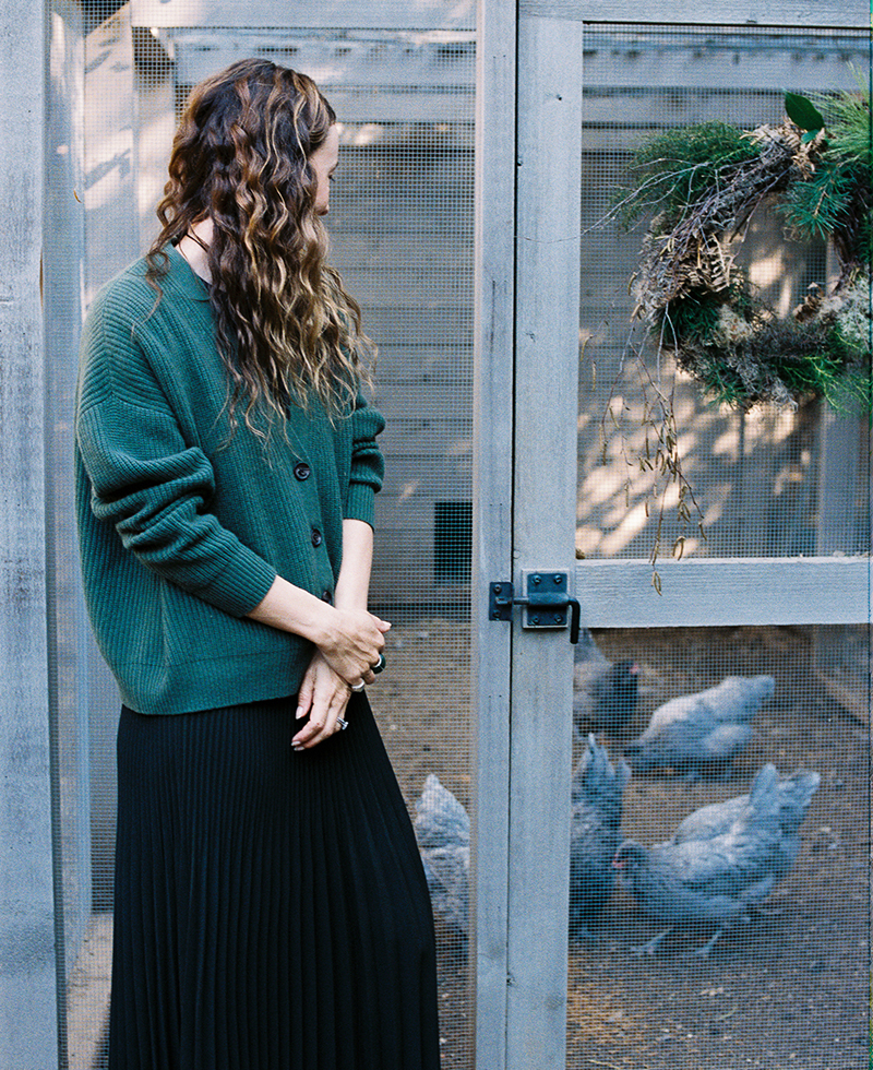 A woman with wavy brown hair, wearing a green cardigan and black skirt, stands outside a chicken coop, looking at several gray chickens inside. A green wreath hangs on the coop door.