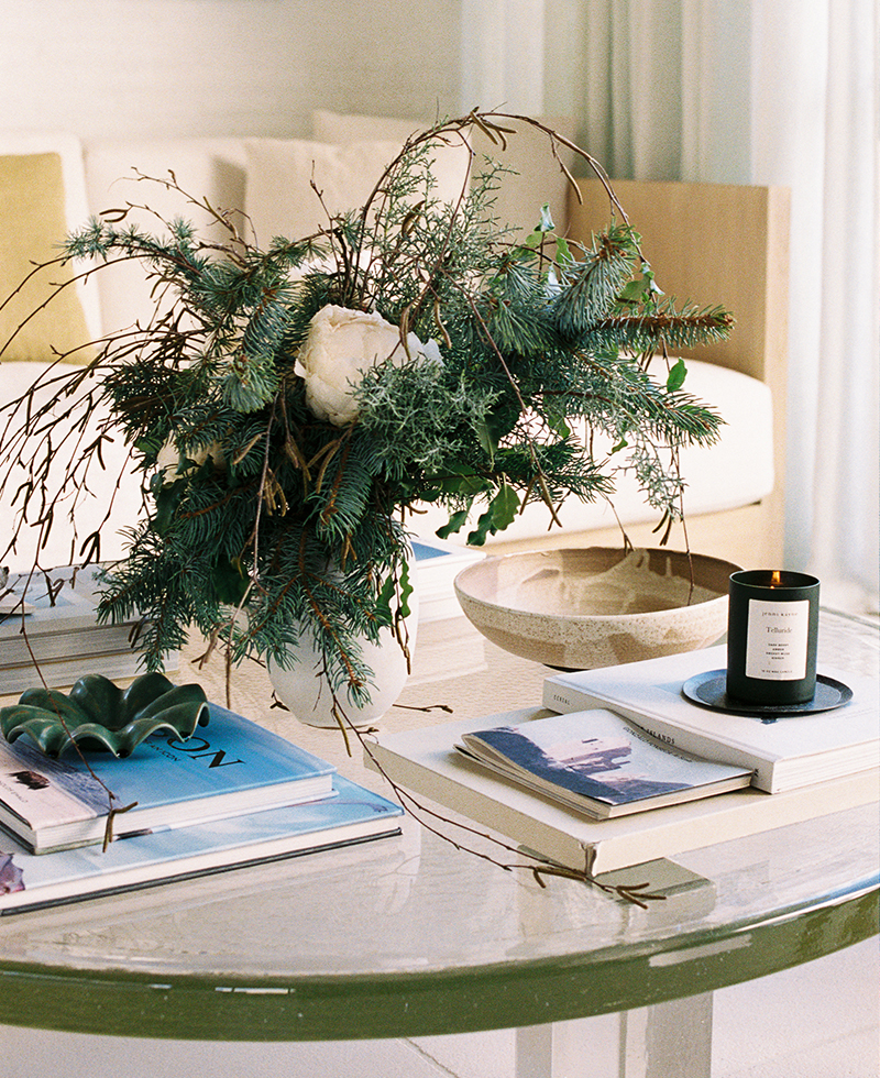 A white round coffee table displays a floral arrangement with greenery and white flowers, a lit black candle on a tray, a beige bowl, and several stacked books. A sofa with light cushions is in the background.