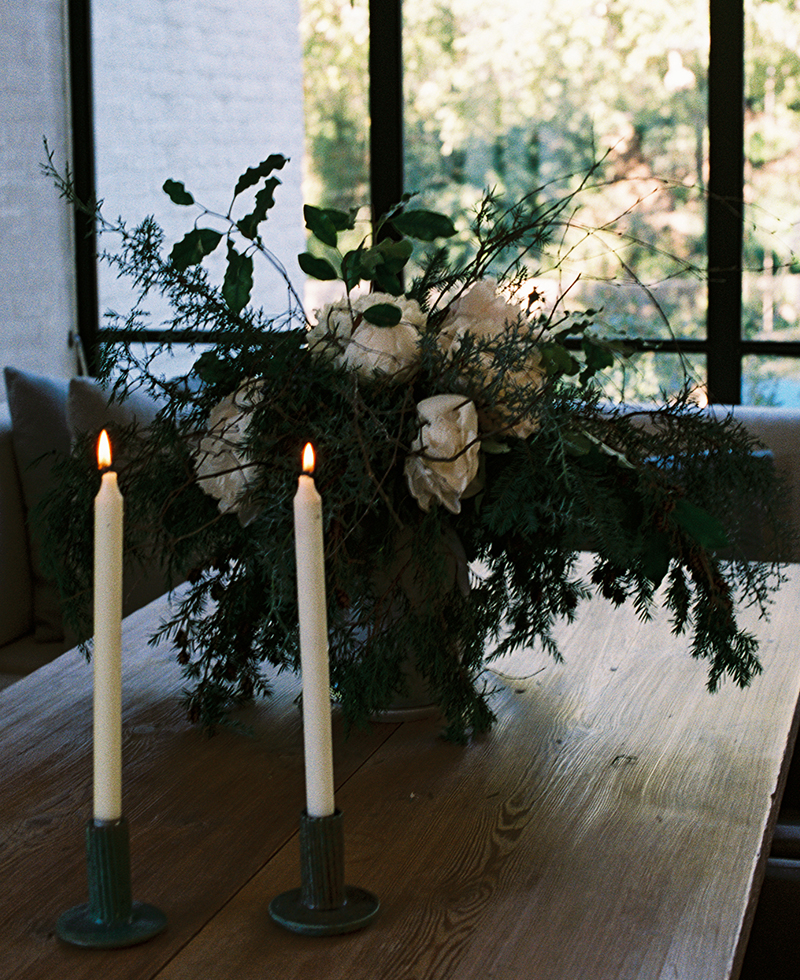 A wooden table with two lit white candles in green holders and a large floral arrangement of white roses and greenery, set by a window with trees visible outside.