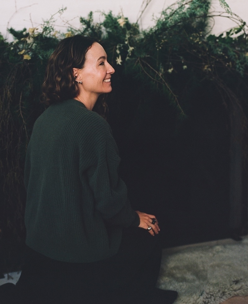 A woman with wavy brown hair, wearing a dark sweater, sits on the floor and smiles, with greenery and plants in the background.