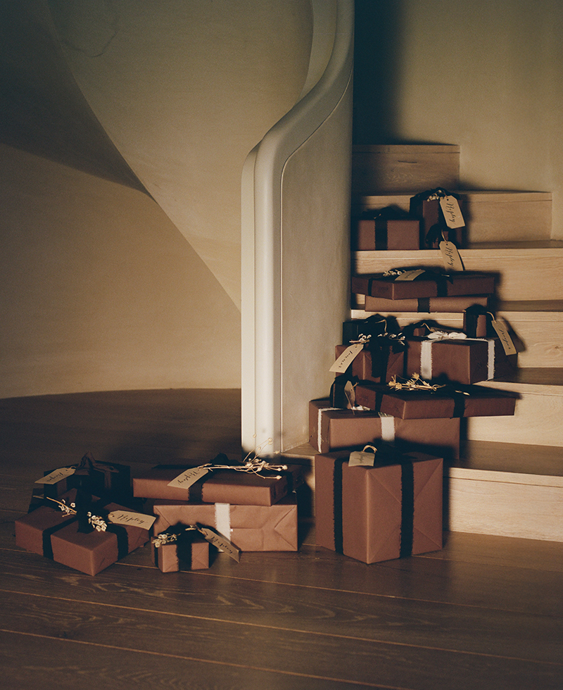 A group of neatly wrapped brown gift boxes with black ribbons and tags are stacked on light wooden stairs and the floor, near a curved white banister in a softly lit room.