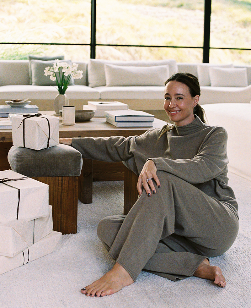 A woman in a cozy, taupe lounge set sits barefoot on a white carpet, smiling and leaning against a wooden table with wrapped gifts, books, and a vase of flowers in a bright, modern living room.