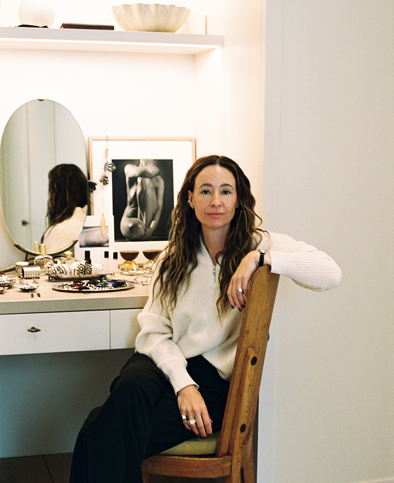 A woman with long wavy hair, wearing a cream sweater and black pants, sits on a wooden chair by a vanity table with a round mirror, jewelry, and framed art in a softly lit room.