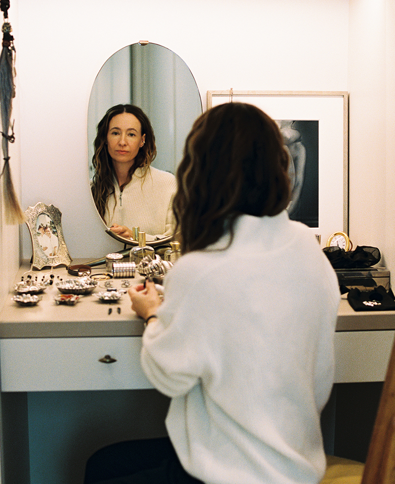 A woman with long brown hair sits at a vanity table, looking at her reflection in an oval mirror. The table is covered with jewelry and beauty items, and framed photos are visible in the background.
