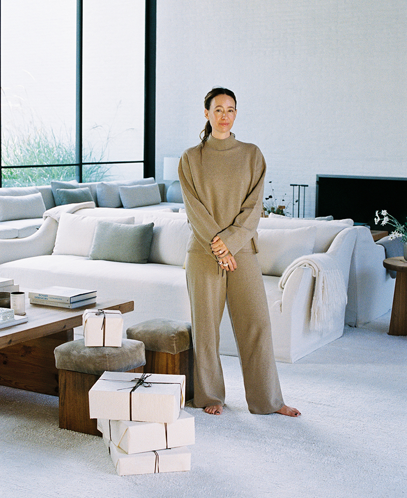 A woman in a beige loungewear set stands barefoot in a modern, light-filled living room with a white sofa and several wrapped gift boxes stacked on the floor nearby.