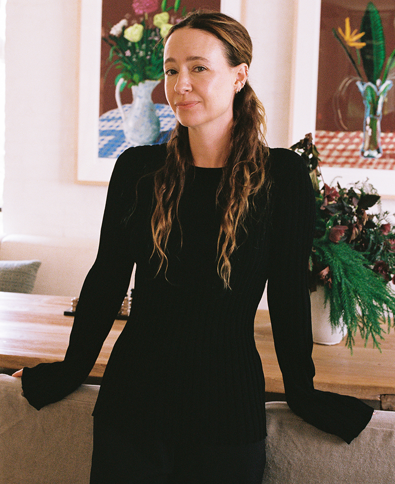 A person with long wavy hair, wearing a black long-sleeved top from their holiday wardrobe, leans against a chair in a well-lit room adorned with floral arrangements and framed art in the background.