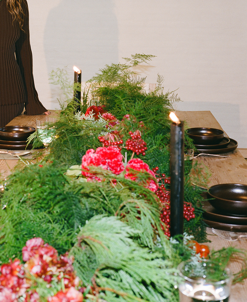A wooden dining table is decorated with lush green foliage, red flowers, and black candlesticks with lit candles. Dark plates are set at each place. A person in a brown outfit stands partially out of frame.