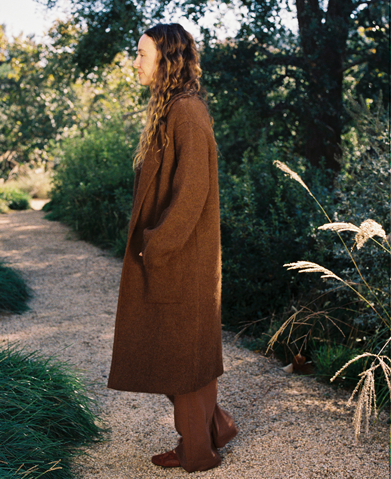 A person with long wavy hair wearing a stylish winter coat and brown pants stands on a gravel path surrounded by greenery and trees.