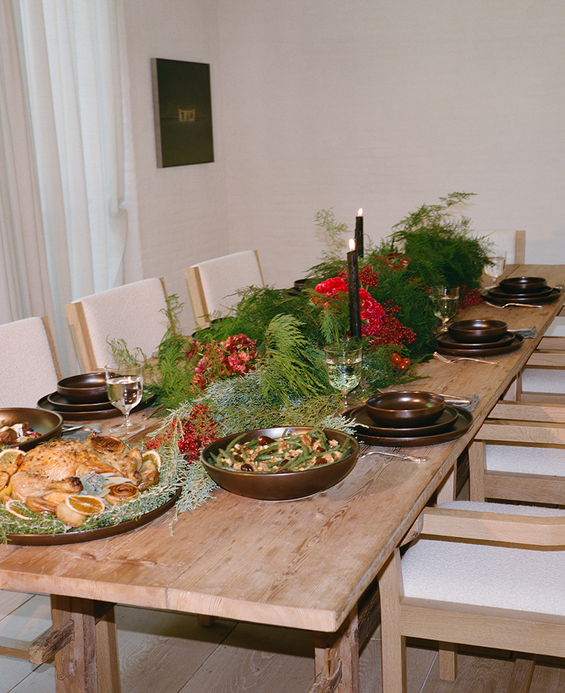 A wooden dining table set for a holiday dinner features brown dishes, glasses of white wine, roast chicken, green beans, and festive greenery with red accents as a centerpiece. Lit black candles add a cozy atmosphere.