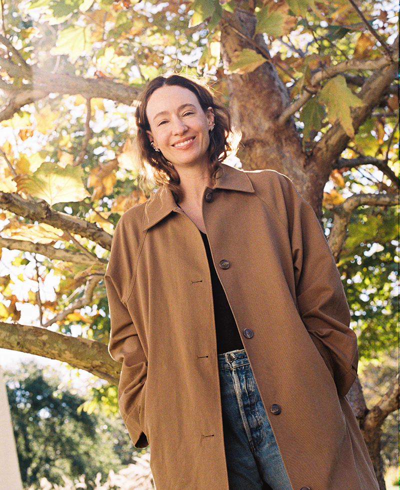 A smiling woman in a brown winter coat stands outdoors in front of a tree with green and yellow leaves, sunlight shining through the branches. She has shoulder-length hair and looks relaxed and happy.